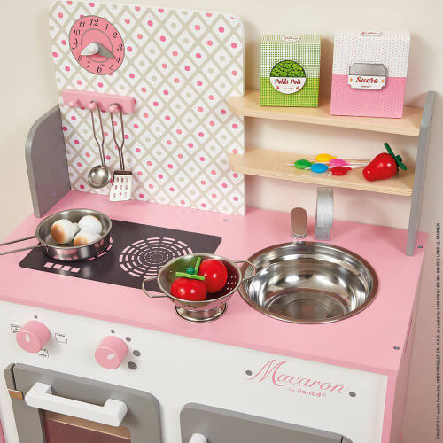 Pink and white wooden toy kitchen with a clock, sink, stove, oven, and play accessories like utensils and food.