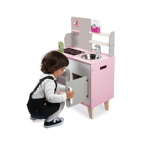 Young child playing with a pink and grey wooden toy kitchen featuring a sink, stovetop, and opening cabinet doors.