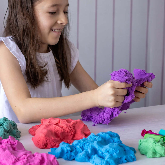 A girl pulls apart purple dough next to mounds of pink, red, blue, and green modeling sand on a white table.