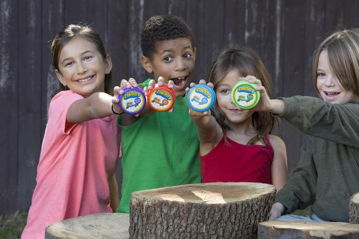 Four smiling children hold up purple, red, blue, and green round pods while sitting behind tree stumps outdoors.