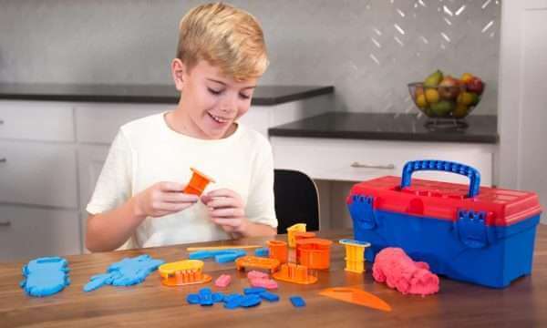 Smiling child plays with pink modeling sand and colorful plastic tools next to a blue and red toy toolbox on a table.