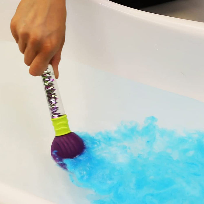 Hand holds a purple toy brush with a glitter-filled handle in white bathwater as a blue bath bomb dissolves.