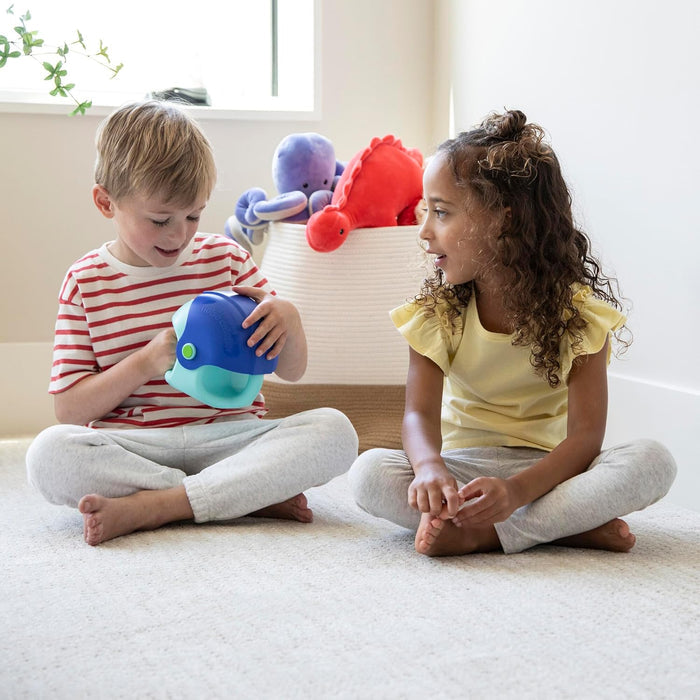 Two children sit on a rug playing with a blue and teal whale-shaped plastic toy.