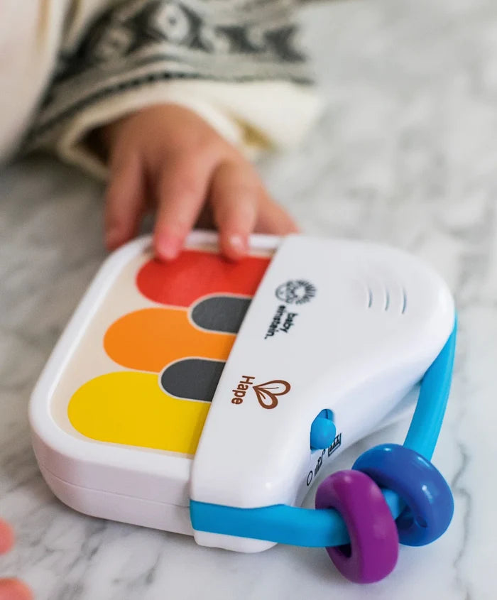 Baby's hand playing a white handheld toy piano with red, orange, and yellow keys and a blue bead handle.