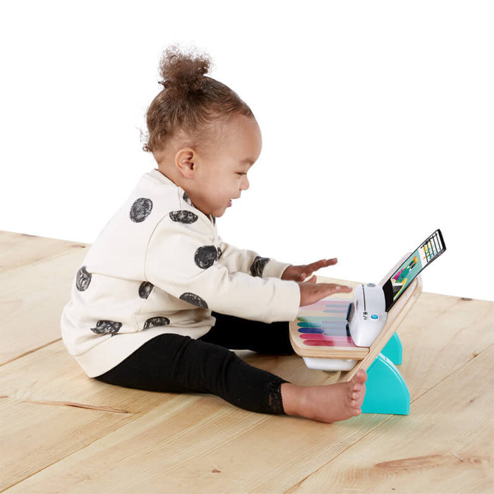 Toddler in a polka dot sweater plays a wooden toy piano with rainbow keys and teal base on a light wood floor.