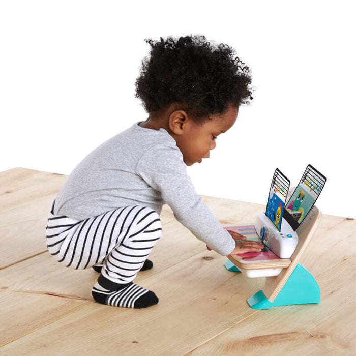 Toddler playing with a white and wooden piano toy featuring rainbow touch keys, teal stand, and sheet music cards.