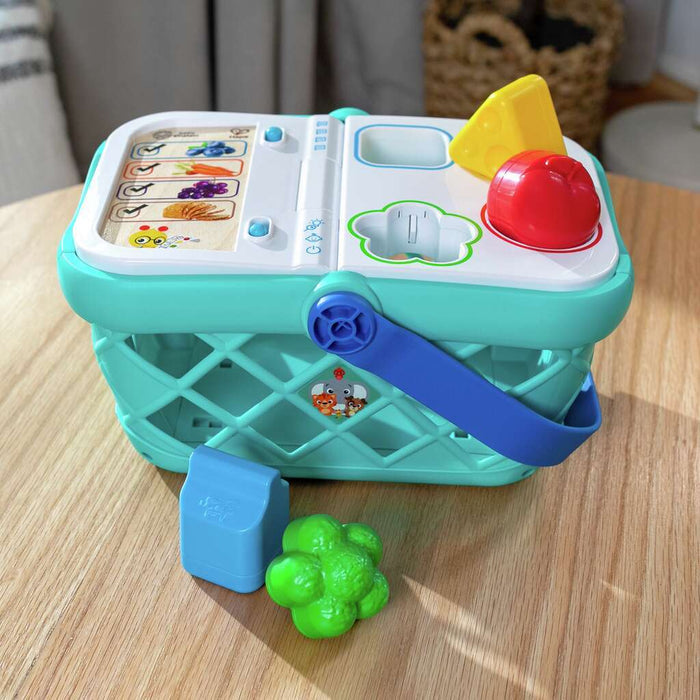 Mint green toy basket with interactive white lid, colorful shape-sorting food, and blue handle on a wooden table.