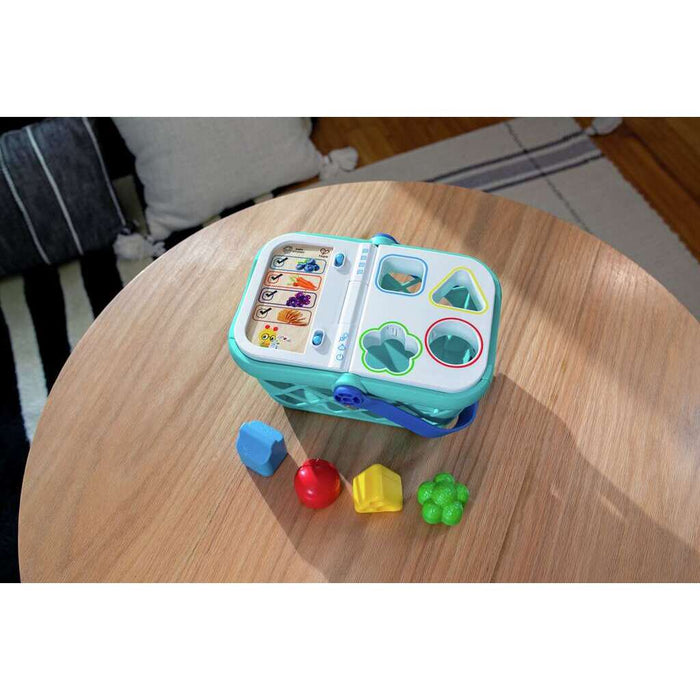 Teal toy shopping basket with a shape-sorting lid and four colorful food blocks on a round wooden table.