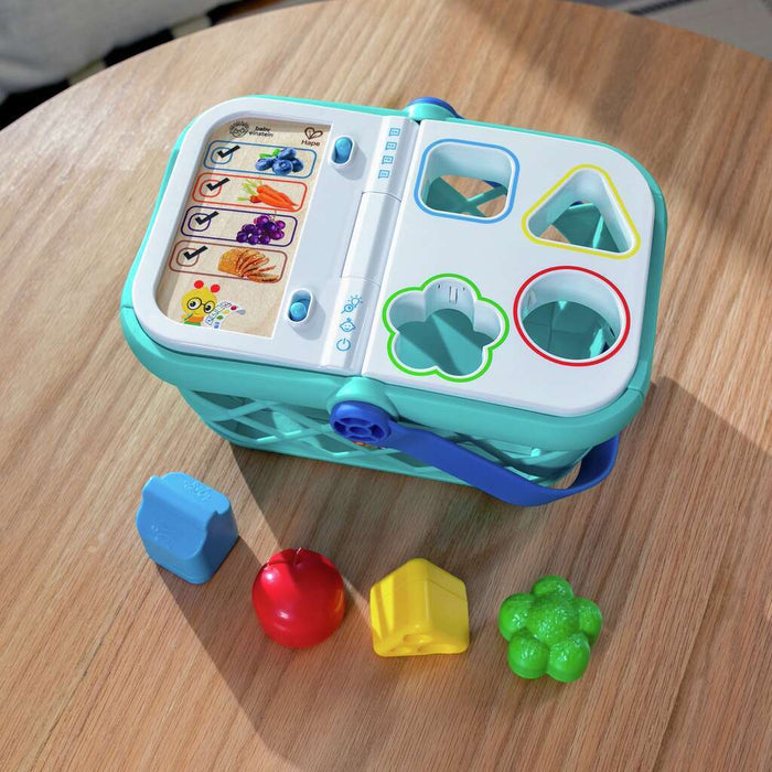 Teal toy shopping basket with a white shape-sorting lid and four colorful food pieces on a wooden table.
