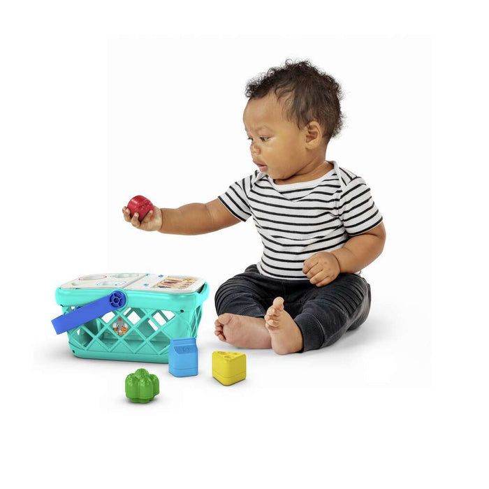 Baby sits by a turquoise shopping basket toy, holding a red apple shape with colorful food blocks nearby.