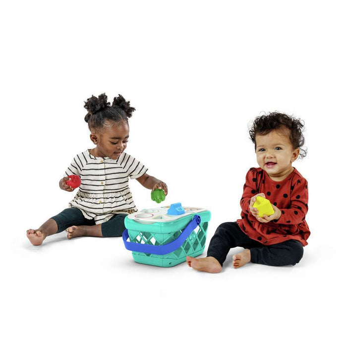 Two toddlers play with a teal toy shopping basket and colorful shape-sorting food pieces on a white background.