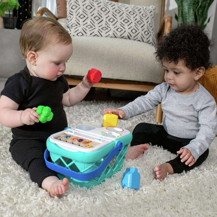 Two infants play with a teal shape-sorting shopping basket and colorful food toys on a white shag rug.