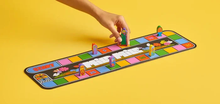 Hand moves a cartoon piece on a long, colorful board game with numbered squares, set against a solid yellow background.