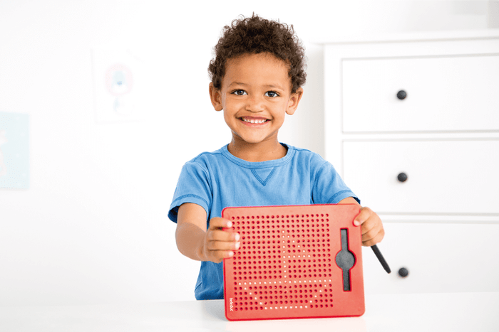 Smiling child holding a red magnetic drawing board with metal beads forming a sailboat and a black stylus.