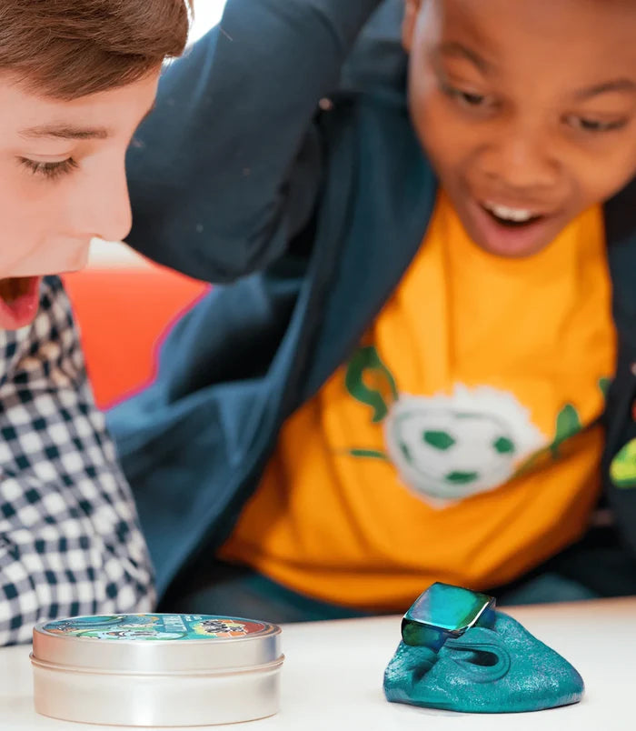 Two boys look at shimmering teal magnetic putty and a metal cube magnet next to a silver tin with a colorful label.