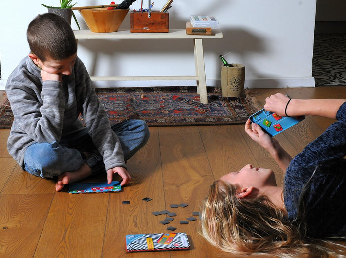 Two children playing with compact magnetic puzzle games in slim, colorful metal travel tins on a wooden floor.