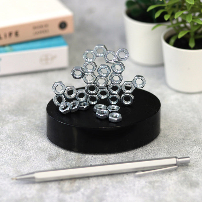 Abstract sculpture of silver hexagonal nuts stacked on a circular black magnetic base, displayed on a desk.