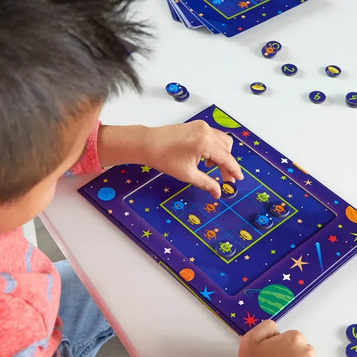 Child's hand placing colorful alien magnets on a purple galaxy-themed 3x3 grid board with star and planet designs.