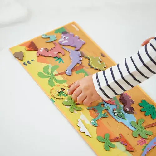 Child's hand arranging colorful magnetic dinosaur shapes on a prehistoric landscape-themed fold-out board.