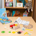 Clock kit with colorful gears, clear casing, and instructions spread on a table next to its blue box and a toy shelf.