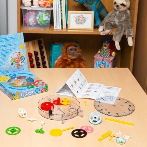 Clock kit with colorful gears, clear casing, and instructions spread on a table next to its blue box and a toy shelf.