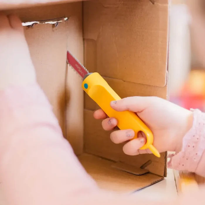 Child's hand holding a yellow plastic hand saw with a serrated blade, cutting into a brown cardboard box.