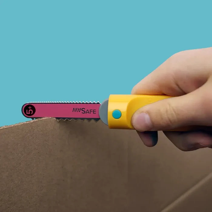 Hand using a yellow saw with a pink serrated blade to cut cardboard against a bright blue background.