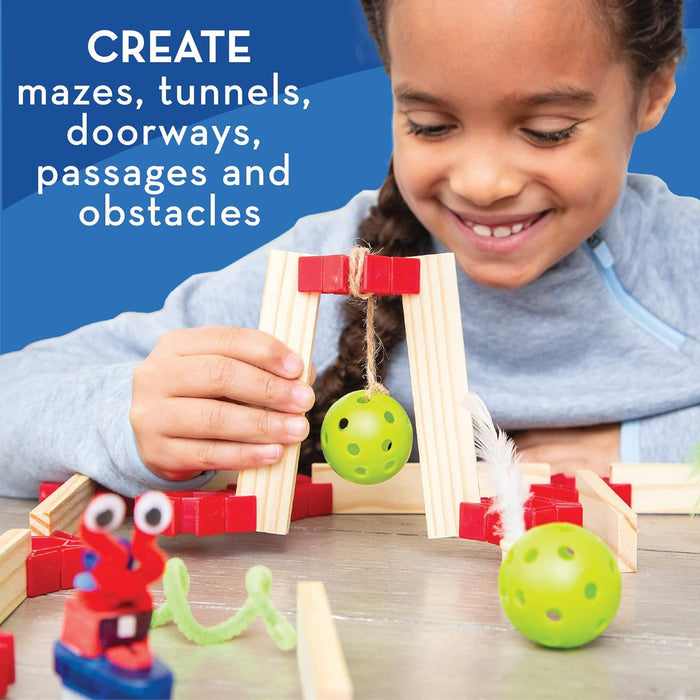 A smiling child builds a maze with wooden planks, red connectors, green balls, and a small toy robot.