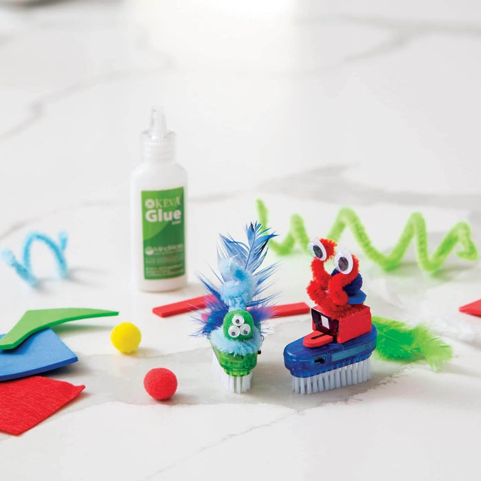 Two colorful brush bots with googly eyes and feathers near glue and craft supplies on a white surface.