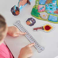 Child's hands point at a bone-shaped number line next to illustrated prehistoric-themed game pieces on a white table.