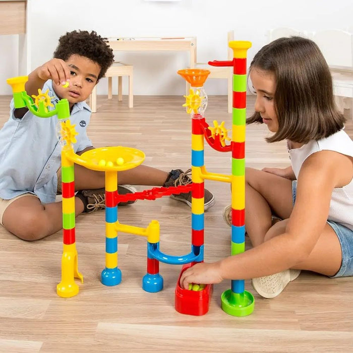 Children playing with a colorful toy tower made of bright plastic tubes, funnels, and spinning gears on a wooden floor.