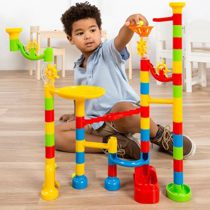 Boy playing with a colorful plastic marble run featuring tall tubes, funnels, and spinning wheels on a wood floor.
