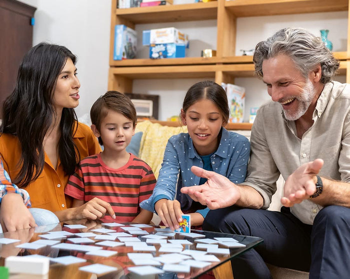 Family playing memory game with blue square tiles on a glass table; a girl flips a card to reveal a colorful hero.