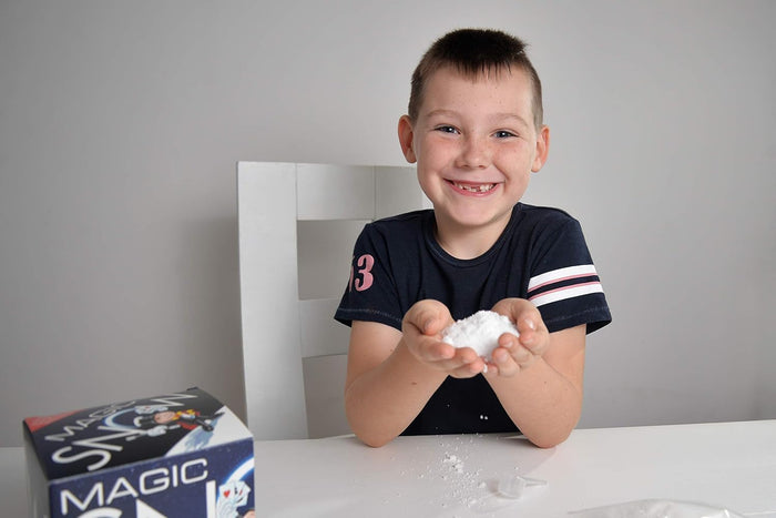 Smiling boy holds a handful of fluffy white magic snow next to the product packaging on a white table.