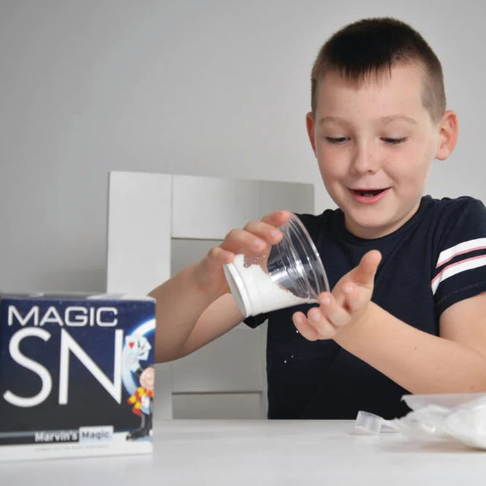 A smiling boy pours white artificial snow from a clear cup into his palm, next to the Magic Snow product box.