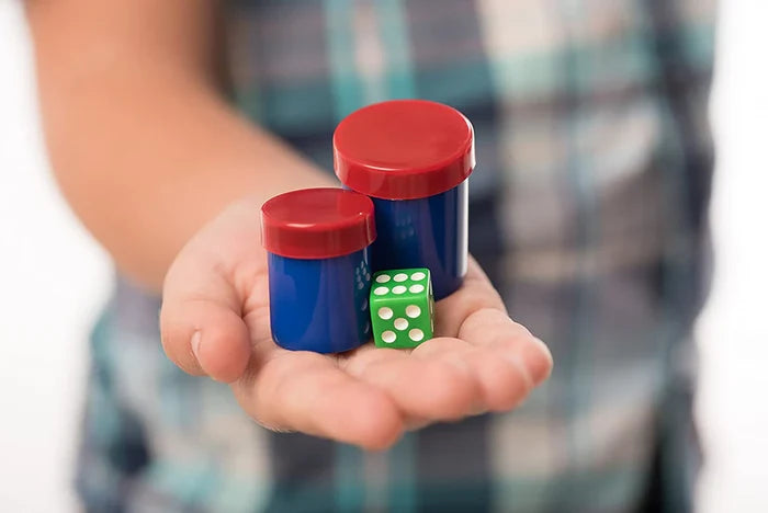 A hand holding two blue cylindrical canisters with red lids and a green six-sided die.