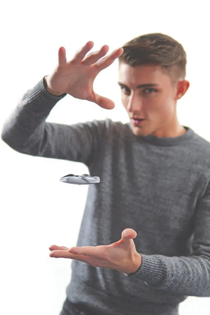 Man in a grey sweater levitating a small silver saucer toy between his hands against a white background.