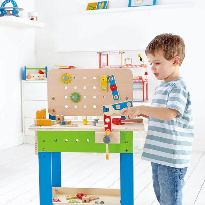 A child building at a wooden toy workbench with a green frame, blue legs, a pegboard, and colorful tools.