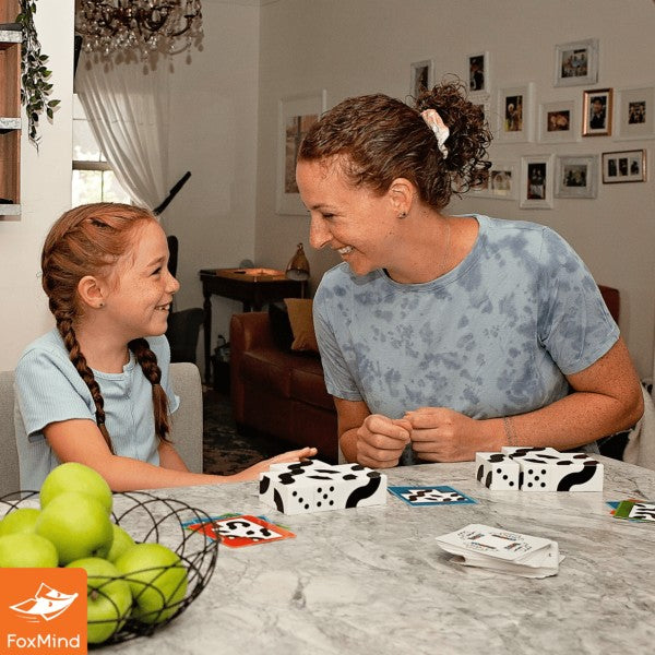 Woman and child play a game with black and white patterned blocks and cards on a marble table with green apples.