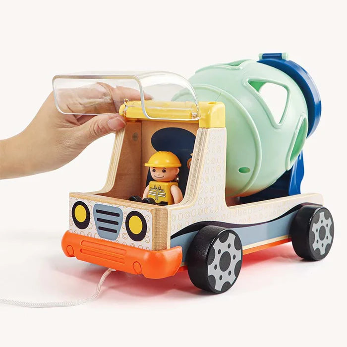 Hand lifting the clear lid of a wooden toy cement truck with a green shape-sorter drum and small driver figure.