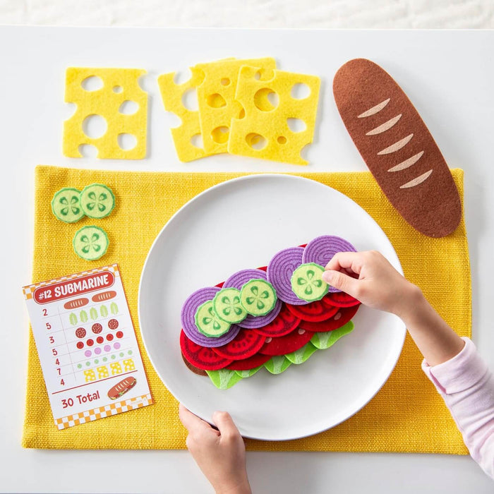 Child's hands assembling a toy sandwich with colorful felt ingredients and a numbered menu card on a white plate.