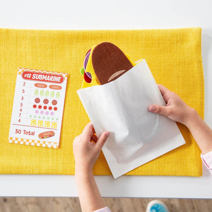 Child places felt sandwich pieces in a white bag next to a counting order card on a yellow textured mat.
