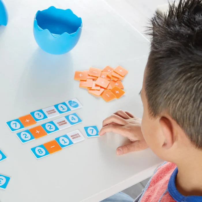 Child arranging blue number and orange operator tiles into math equations on a white table next to a blue egg container.