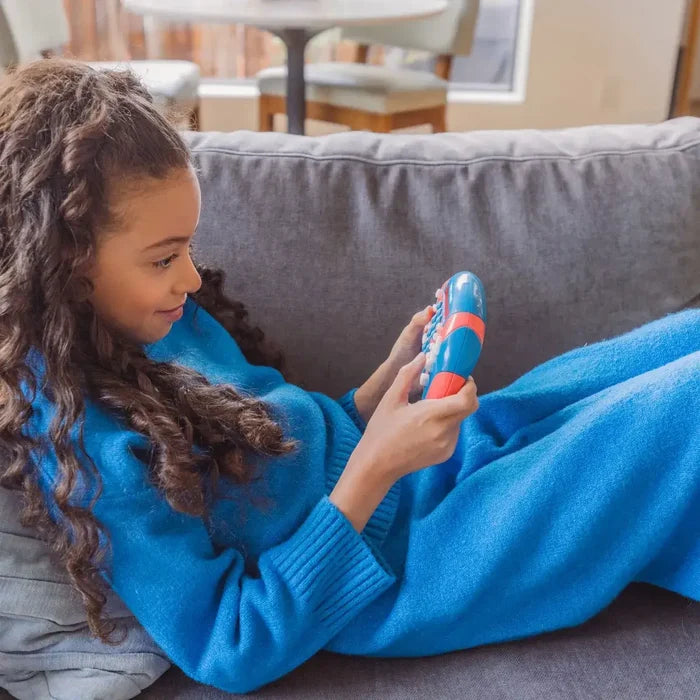 Young girl on a grey sofa playing with a blue and red handheld electronic math game with white buttons.