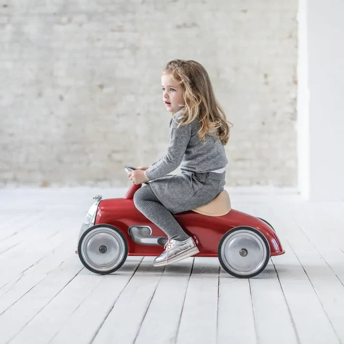 Young child on a vintage-style red ride-on car with silver wheels and a tan seat against a white brick wall.