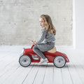 Young child on a vintage-style red ride-on car with silver wheels and a tan seat against a white brick wall.