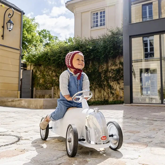A child sits on a white, vintage-style ride-on car with a chrome grille and steering wheel on a sunny stone path.