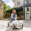 A child sits on a white, vintage-style ride-on car with a chrome grille and steering wheel on a sunny stone path.