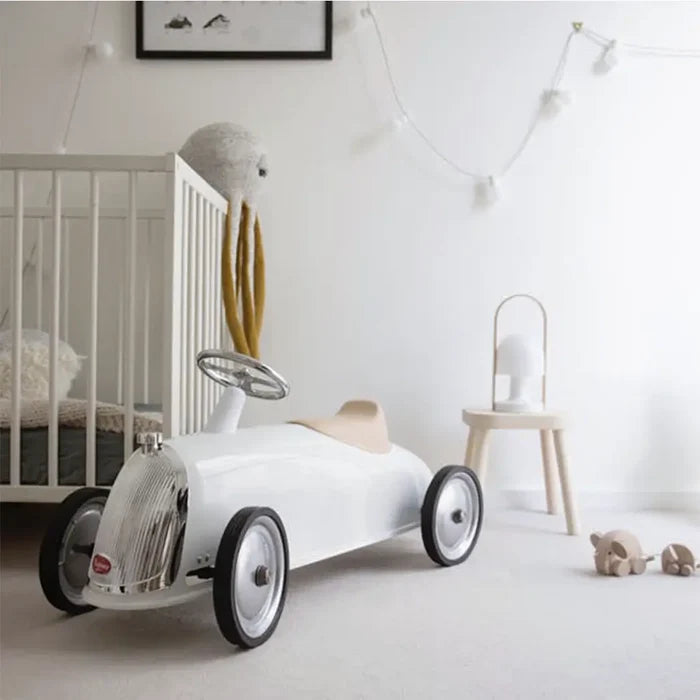White vintage-style ride-on car with chrome grille, silver steering wheel, and beige seat in a modern nursery.