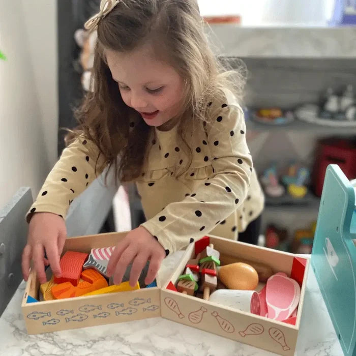 Young girl playing with wooden crates filled with colorful toy fish, meat skewers, steak, and a chicken leg.